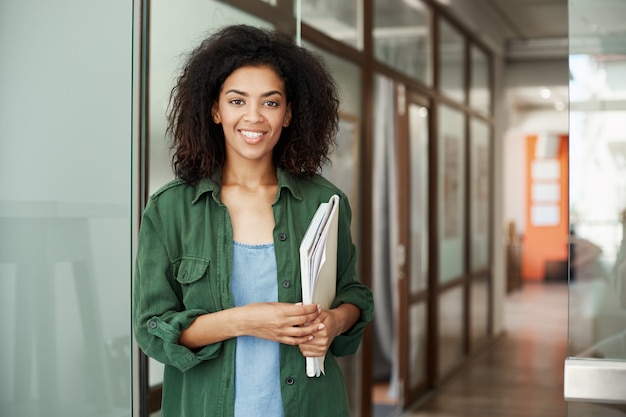 Estudiante de mujer hermoso africano alegre que sonríe sosteniendo los libros en universidad. Concepto de educación