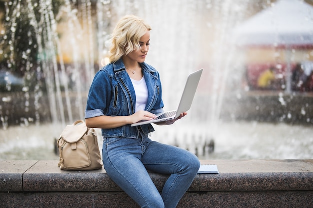 Estudiante de mujer rubia ocupada trabaja en su computadora portátil cerca de la fuente en la ciudad en el día