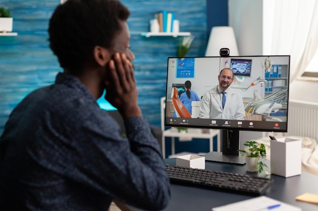 Estudiante negro con dolor de muelas discutiendo el tratamiento médico con el médico durante la conferencia de la reunión por videollamada en línea. Hombre que sufre una enfermedad dolorosa. Llamada de videoconferencia de telesalud en computadora