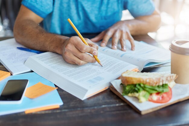 estudiante negro que subraya información importante en un libro de texto con lápiz mientras hace una investigación de historia en el comedor de la universidad durante el almuerzo; teléfono, café y comida descansando sobre la mesa