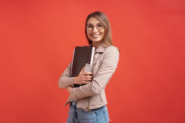 Estudiante o pasante sonriente joven en las lentes que se colocan con una carpeta en la pared roja.