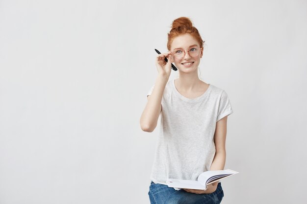 Estudiante pelirroja sonriente corrigiendo gafas sosteniendo el cuaderno.