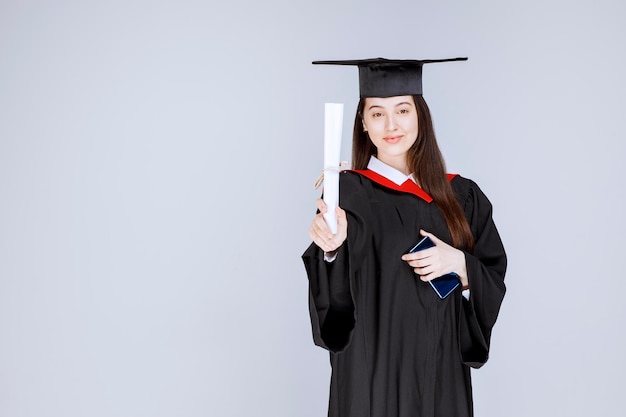 Estudiante de posgrado en bata teléfono móvil en mano sosteniendo diploma. Foto de alta calidad