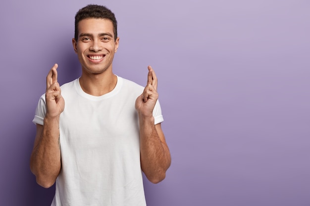 Un estudiante que ora levanta los dedos cruzados, pide éxito y buena suerte, sonríe positivamente, ora antes de aprobar un examen importante en su vida, viste una camiseta blanca informal, posa sobre una pared violeta