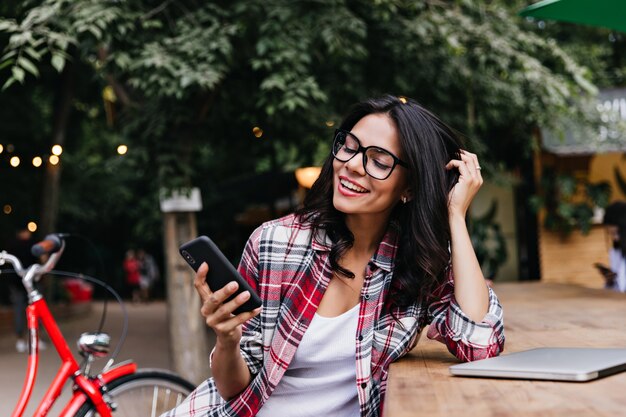 Estudiante relajado sentado en la calle con ordenador portátil y teléfono. Encantadora chica latina con cabello negro posando junto a su bicicleta.