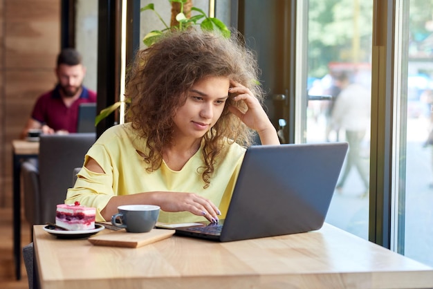 Estudiante rizado que estudia trabajar con una computadora portátil sentada en un café