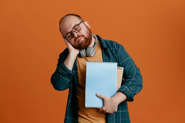 Un estudiante con ropa informal que usa anteojos con auriculares que sostienen cuadernos que se ven cansados y con exceso de trabajo quiere dormir con la cabeza apoyada en la palma de la mano sobre un fondo naranja