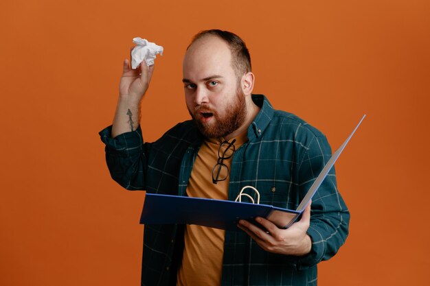 Foto gratuita estudiante con ropa informal sosteniendo una carpeta de oficina con papel arrugado que parece sorprendido de pie sobre un fondo naranja