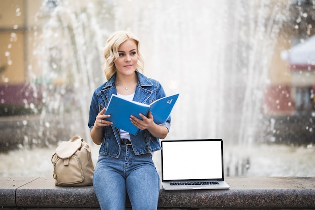 Estudiante rubia chica trabaja en su computadora portátil y lee un libro cerca de la fuente en la ciudad en el día