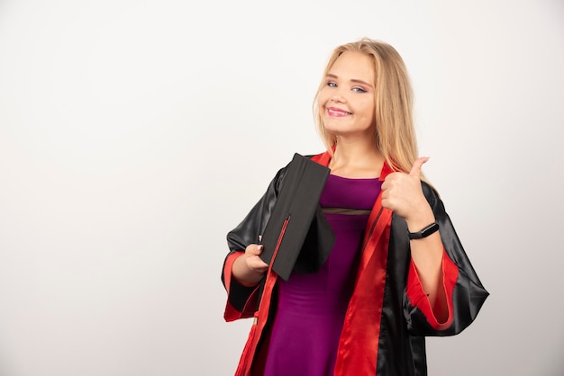 Estudiante rubia sosteniendo su gorra y haciendo pulgares para arriba en blanco.