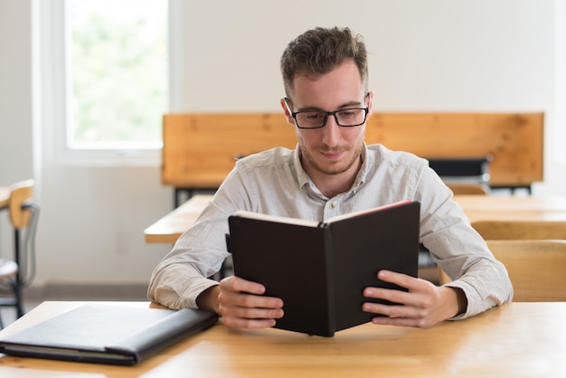 Estudiante serio que lee el libro de texto en el escritorio en sala de clase