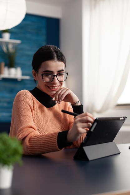 Estudiante sonriente con gafas que sostienen la pantalla táctil de la tableta. Freelancer trabajando a distancia interactuando con tablet-pc. Chica universitaria en el estudio mirando casualmente la pantalla mientras sostiene el lápiz táctil.