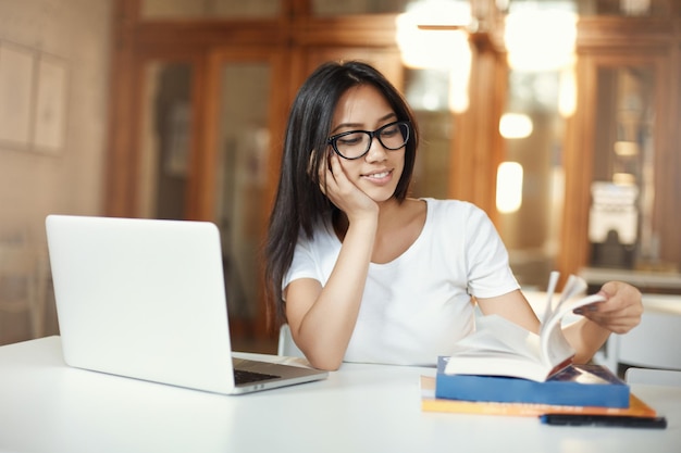 Estudiante sonriente hojeando el libro de estudio mientras trabaja en su diploma de licenciatura en una biblioteca de espacio abierto