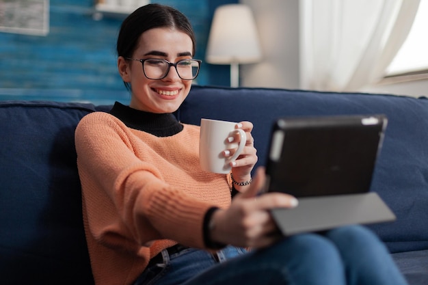 Estudiante sonriente en el sofá hablando casualmente en una videollamada usando una tableta. Creador de contenido con gafas en el sofá viendo videos en las redes sociales. Influenciador digital relajándose con una taza de café o té.