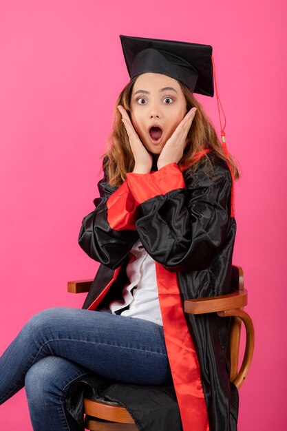Estudiante sorprendido vistiendo toga de graduación y sentado en una silla.