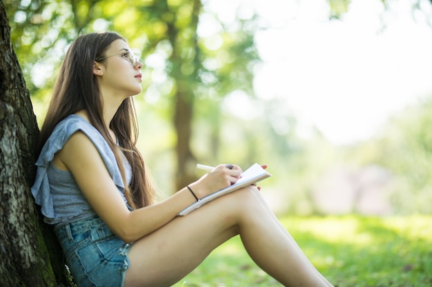 Estudiante tomando notas en su cuaderno. Mujer sonriente joven sentada en el parque haciendo asignaciones. Vida en el campus, educación, concepto de inspiración