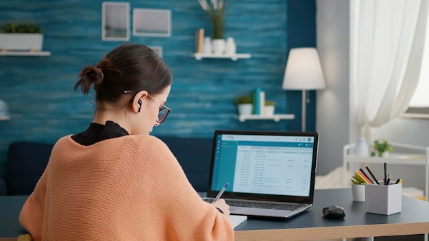 Estudiante trabajando desde casa en una computadora portátil para hacer investigación en Internet, aprendiendo educación universitaria en clases en línea. Mujer haciendo tareas en el cuaderno, tomando notas y aprendiendo.