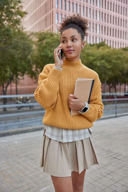 estudiante en traje elegante habla por teléfono móvil sostiene libro de texto mira hacia otro lado tiene conversación durante el descanso posa contra edificios