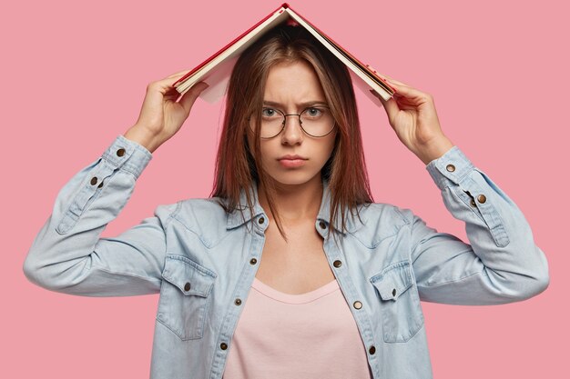 Una estudiante universitaria europea hermosa y seria sostiene un libro sobre la cabeza, se siente cansada de aprender material para el examen, tiene expresión desconcertada, no quiere estudiar, aislado sobre una pared rosa.