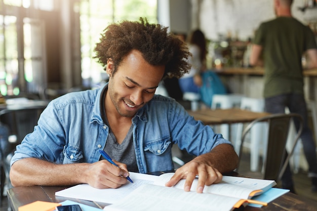 Estudiante universitario afroamericano alegre atractivo que trabaja en la asignación de casa en la cafetería, escribiendo composición o haciendo investigación, con mirada feliz y entusiasta. Personas, conocimiento y educación