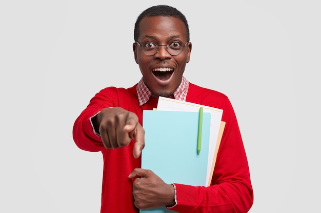 El estudiante universitario afroamericano alegre señala con los dedos índices al frente, tiene una expresión feliz, tiene la piel oscura, vestido con un suéter rojo