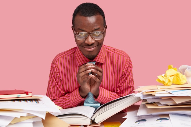 Foto gratuita estudiante universitario afroamericano complacido de piel oscura mantiene la mano en un gesto intrigante, tiene una buena idea para expresar, lee un libro grueso