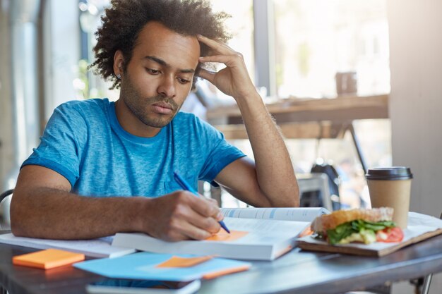 Estudiante universitario afroamericano serio concentrado con barba haciendo tareas en casa, preparándose para la lección de español, escribiendo nuevas palabras del texto en notas adhesivas durante el desayuno en la cafetería