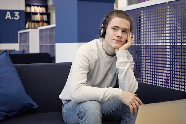 Estudiante universitario guapo en suéter y jeans usando auriculares inalámbricos mientras escucha información, preparándose para la prueba en la universidad