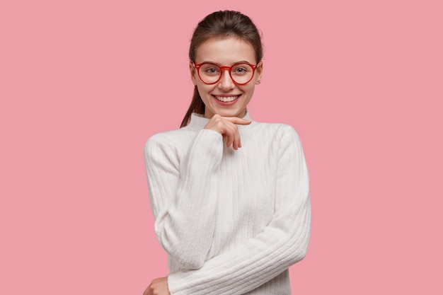 Estudiante universitario joven positivo viste suéter blanco de invierno, gafas de borde rojo, mantiene la mano debajo de la barbilla, sonríe ampliamente