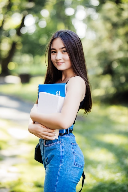 Estudiante universitario con libros caminando en el campus yendo a clase sonriendo. Muchacha asiática multirracial sonriente joven de la mujer con el retrato al aire libre del bolso.