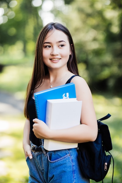 Estudiante universitario con libros caminando en el campus yendo a clase sonriendo. Muchacha asiática multirracial sonriente joven de la mujer con el retrato al aire libre del bolso.