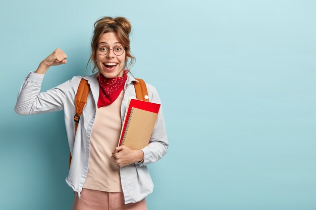 Estudiante universitario optimista y feliz con apariencia europea, levanta los puños cerrados, muestra músculos, celebra el año escolar terminado con éxito