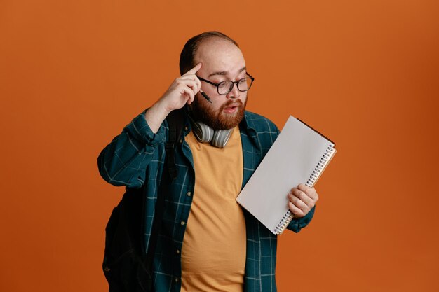 Estudiante vestido con ropa informal y gafas con auriculares y mochila sosteniendo un cuaderno y un bolígrafo que parece desconcertado apuntando con el dedo índice a su sien de pie sobre un fondo naranja
