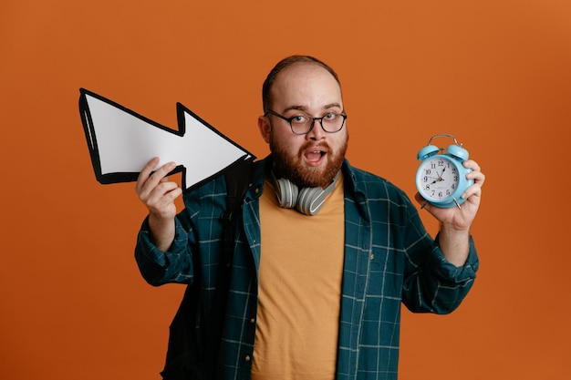 Foto gratuita estudiante vestido con ropa informal con gafas y auriculares sosteniendo un despertador y una flecha mirando a la cámara feliz y positivo sonriendo alegremente de pie sobre un fondo naranja