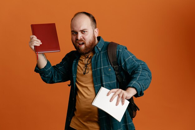 Estudiante vestido con ropa informal con mochila sosteniendo cuadernos que parece molesto parado sobre fondo naranja
