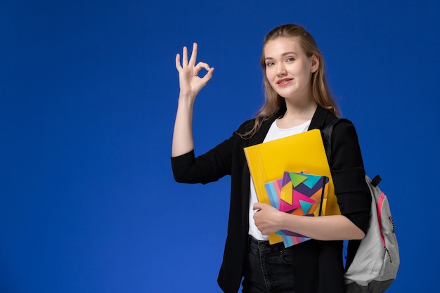 Estudiante de vista frontal en camisa blanca y chaqueta negra con mochila sosteniendo archivos con cuadernos en la lección de la universidad de la universidad de la pared azul