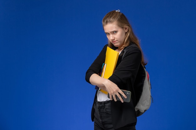Estudiante de vista frontal en chaqueta negra con mochila sosteniendo el archivo y el cuaderno con el teléfono en la pared azul lecciones college university