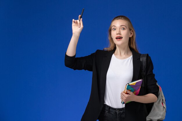 Estudiante de vista frontal en chaqueta negra con mochila sosteniendo bolígrafo y cuaderno en libros de pared azul lecciones de la universidad de la universidad