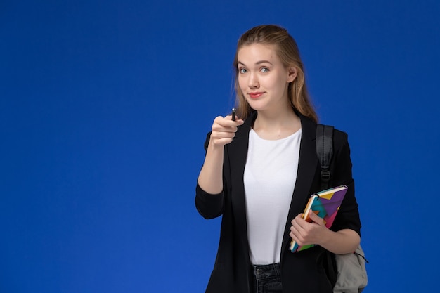 Estudiante de vista frontal en chaqueta negra con mochila sosteniendo bolígrafo y cuaderno en la pared azul lecciones de la universidad de la universidad