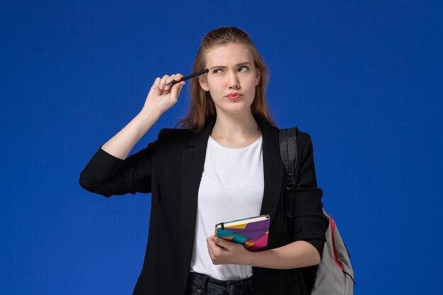 Estudiante de vista frontal en chaqueta negra con mochila sosteniendo bolígrafo y cuaderno pensando en libros de pared azul escuela universidad lección