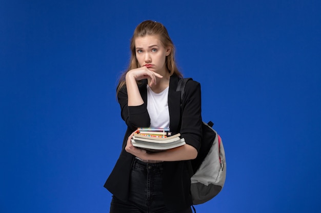 Estudiante de vista frontal en chaqueta negra con mochila sosteniendo libros en la escuela de arte de dibujo de pared azul claro