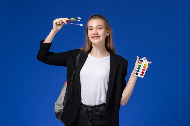 Estudiante de vista frontal en chaqueta negra con mochila sosteniendo pinturas para dibujar y borla en la pared azul dibujo colegio de la escuela de arte