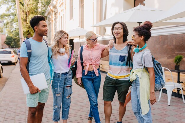 Los estudiantes contentos escuchan a un amigo asiático con gafas que cuenta un nuevo chiste. Las niñas usan jeans y auriculares de moda pasar tiempo con sus compañeros en la calle al lado de la cafetería.