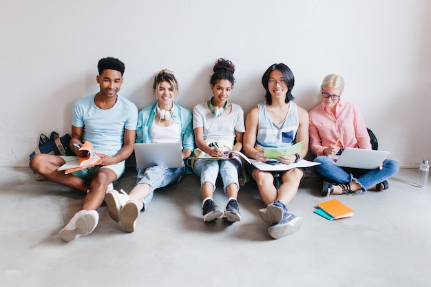 Estudiantes emocionados con computadoras portátiles y libros de texto preparándose para la prueba sentados en el piso. Retrato interior de amigos internacionales que estudian juntos antes de los exámenes.