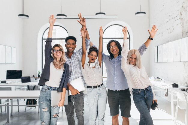 Estudiantes internacionales cansados celebrando el final de los exámenes y riendo en la sala de conferencias. Felices programadores independientes realizados con proyecto largo y posando con una sonrisa, sosteniendo portátiles.