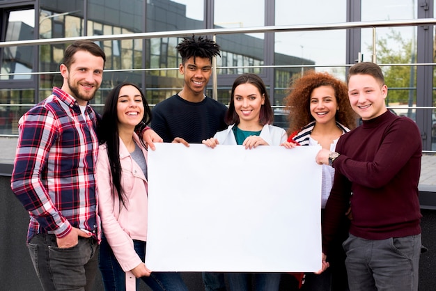 Estudiantes multiétnicos sonrientes que sostienen el cartel blanco en blanco que se coloca delante del edificio de cristal