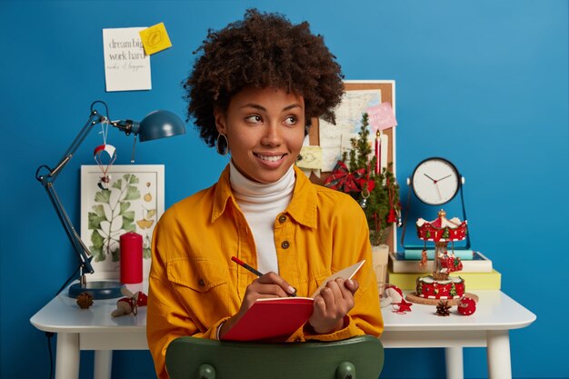 Estudiar, horario de invierno, concepto de vacaciones. Mujer pensativa satisfecha con cabello afro escribe notas en el bloc de notas rojo, hace una lista para hacer antes de la víspera de Navidad