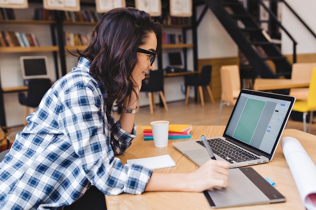 Estudiar momentos de mujer joven morena muy alegre con gafas negras trabajando con el portátil en la mesa en la biblioteca. Estado de ánimo alegre, sonriente, diseño, trabajo creativo, estudiante inteligente, éxito.