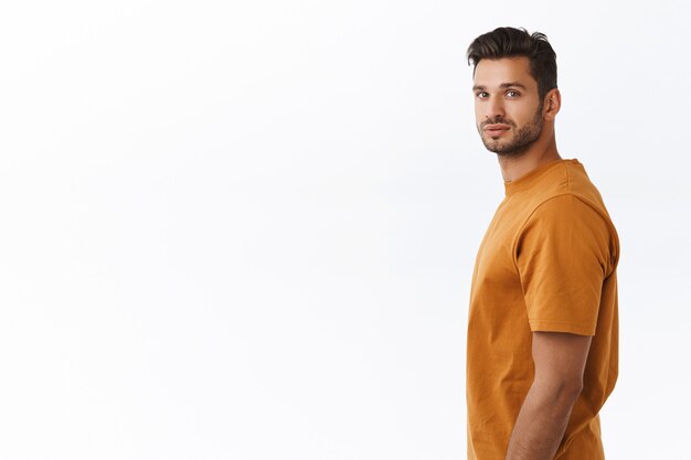 Estudio de disparo chico carismático hipster con barba en camiseta marrón, de pie en el perfil del lado izquierdo, girando la cabeza hacia la cámara y sonriendo, ver algo interesante, prestar atención a una buena promoción