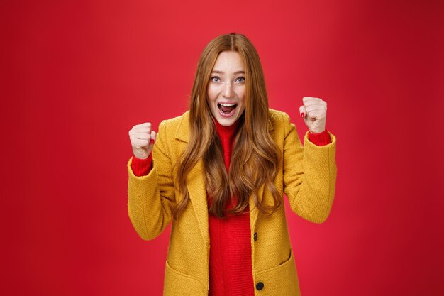 Estudio shto de alegre excitada y guapa chica de jengibre con abrigo amarillo levantando los puños cerrados de alegría y felicidad, triunfando gritando sí desde el éxito y el triunfo, celebrando la victoria sobre la pared roja.